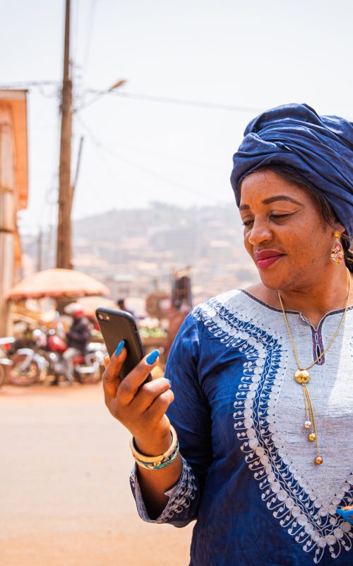An African woman uses her smartphone while she is in the city center, she wears traditional african clothes. An African woman uses her smartphone while she is in the city center, she wears traditional african clothes