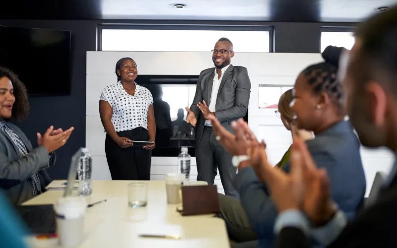 Attractive black businessman being encouraged by diverse multi-ethnic group of coworkers during presentation in office Group of mixed race colleagues in modern meeting room with laptop computer encouraging two attractive African American business professionals leading a collaborative discussion